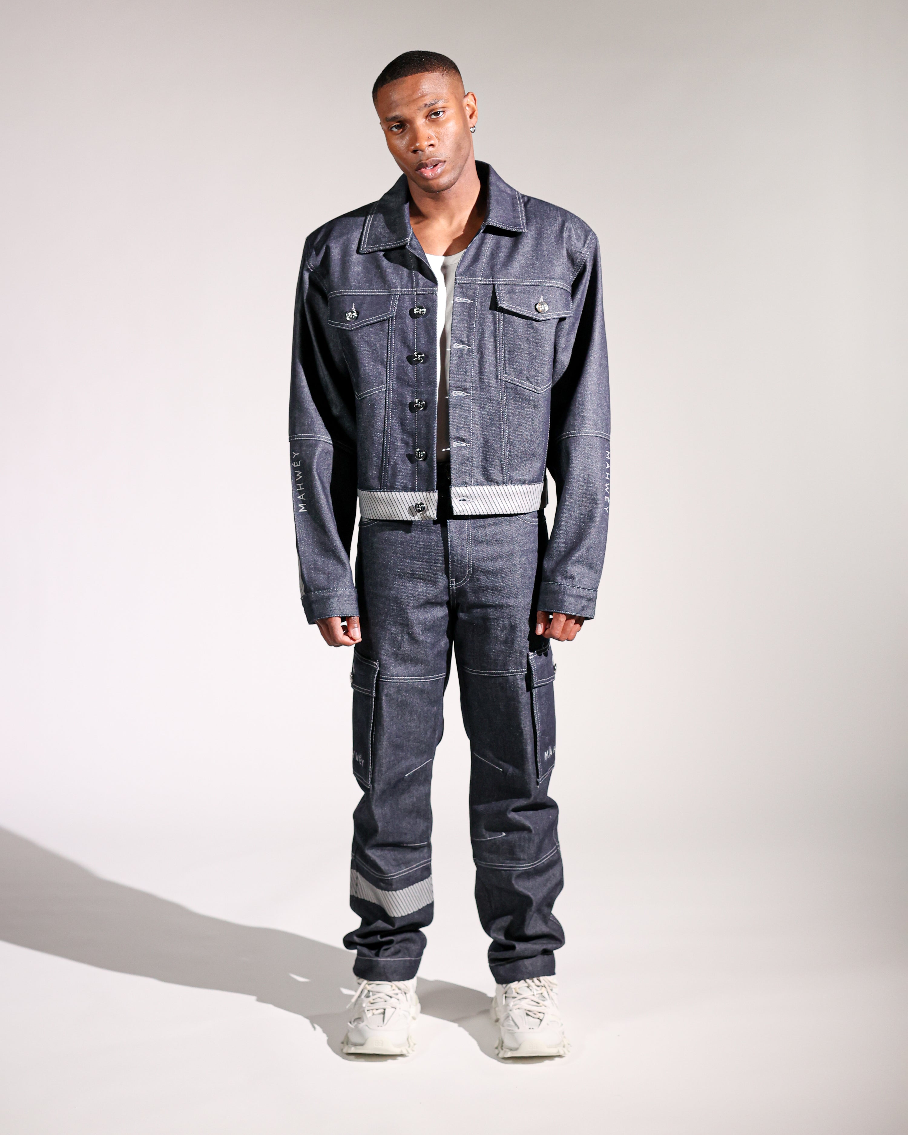 A male model wearing a full-length denim outfit, consisting of a denim jacket and oversized jeans, paired with white sneakers.