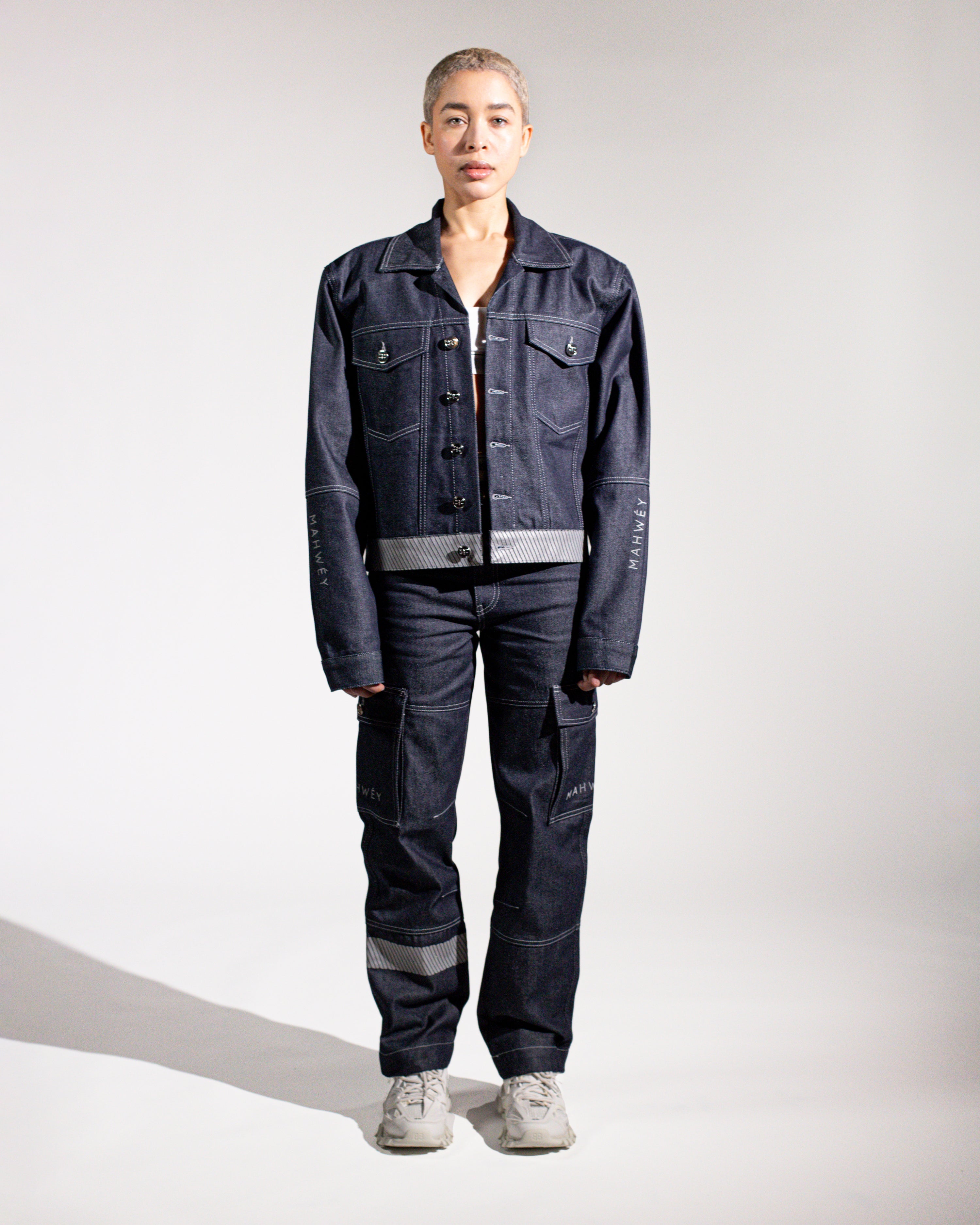 A person standing against a white background, wearing a full-length navy blue outfit consisting of a jacket and pants, paired with white shoes.